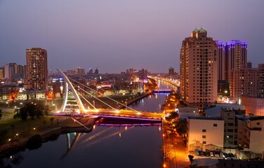 Aerial night skyline of residential buildings by the riverside, with city lights dazzling at blue dusk and the landmark cable-stayed bridge (新臨安橋) spanning the canal, in Tainan City, Taiwan, Asia