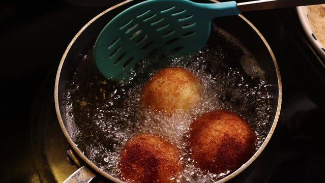 Hand turns Italian arancini rice balls that are frying in hot bubbling oil with spatula.