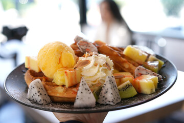 Chocolate waffle with ice cream and whipped cream and chocolate sauce topping watering from top by small jar. Sweet cold European strawberry, kiwi dessert on white plate