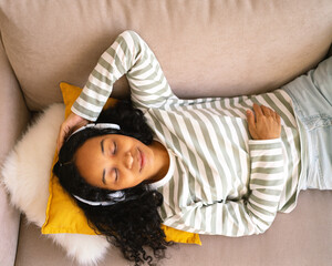 Calm african-american female lying on couch, listening to music in headset. Enjoying audio playing