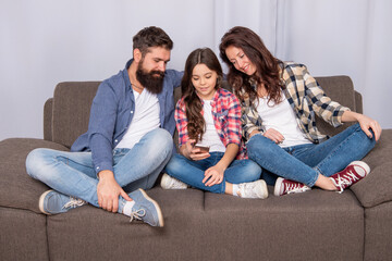 Happy father and mother look at girl child chatting in smartphone sitting on sofa, chat