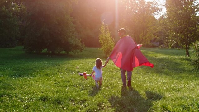 Perfect sunny day for a dad and his small daughter playing together in the middle of the park they walking through the grass the dad wearing a superhero suit