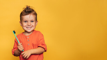 Happy baby toddler boy brushing his teeth with a toothbrush on a yellow background. A place for...
