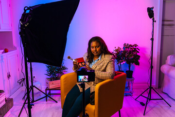 Curvy african american female vlogger in stylish outfit sitting in armchair and applying makeup while filming video for social media on camera on tripod in home studio with bright illumination 