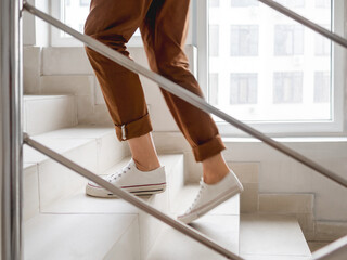Woman in white sneakers and khaki trousers goes upstairs to her apartment. White staircase in apartment building. Casual outfit, urban fashion. Physical exercises.