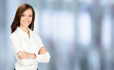 Portrait of smiling businesswoman in crossed arms pose, confident white clothing. Brunette business woman, indoor. Blurred modern office interior background.