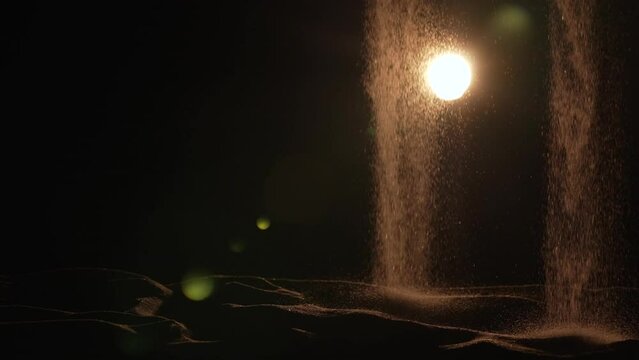 Particles of dry sand are pouring down on black studio background with backlight. Close up shot stream of flowing natural sand grains. Sand flows down in dark. Concept of the flow of life. Slow motion
