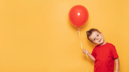 A happy Caucasian boy isolated on a bright yellow background holds a red balloon in his hands. A place for your text or advertisement.