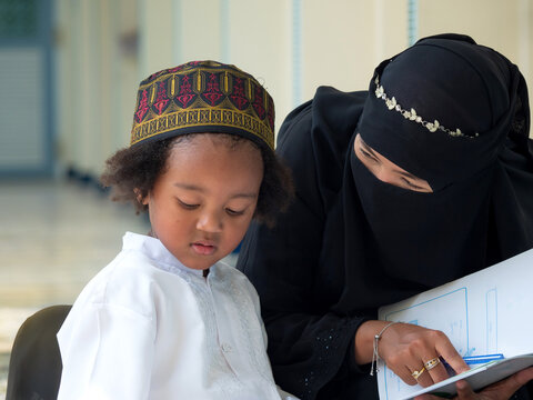 Asian Muslim woman wearing black traditional clothing with hihab and decorated headpiece trying to teach cute little boy to read or pay attention to the book. Mother doing homeschooling.