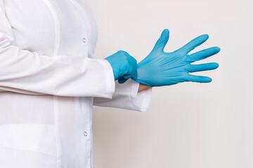 Cropped shot of a young female doctor in a white coat puts on blue medical gloves before examining the patient isolated on a gray background