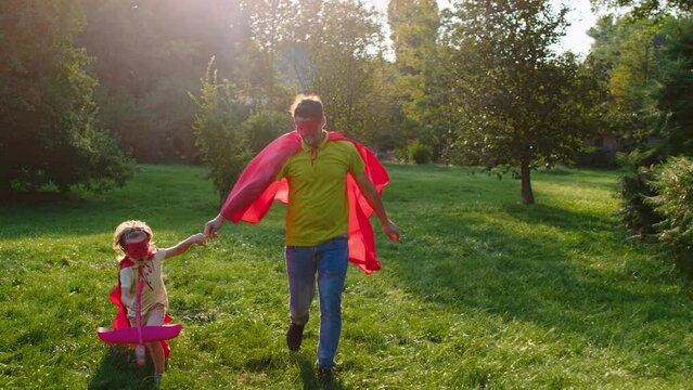 Charismatic and happy dad with his cute small son running in front of the camera with a superhero s suits in the middle of the park while holding hands
