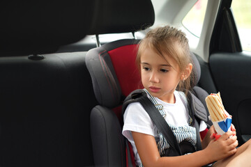 A child in a child's car seat in the back seat, fastened with seat belts, holds a bun with a...