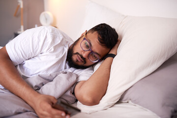 A man wearing spectacles looking at his mobile phone while lying in bed