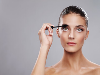 Making her eyes look beautiful. Studio shot of a beautiful young woman applying makeup against a gray background.