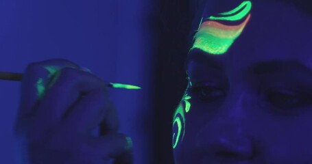 Makeup artist draws on face of young woman with neon paint. Bright colors glowing at ultraviolet illumination of dark club. Thematic party closeup