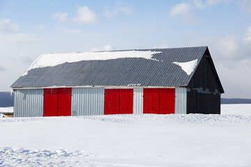 Large metal barn with bright red doors in pristine snowy field against light white clouds and blue sky seen during a winter afternoon, Quebec City, Quebec, Canada