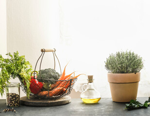 Healthy colorful vegetables in metal basket, olive oil, potted thyme and kitchen utensils on grey kitchen counter at window background with natural light. Plastic free groceries shopping. Front view.