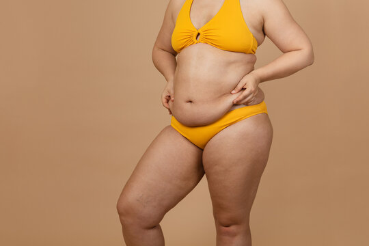 Cropped image of overweight woman, tucking, hiding fat naked big excessive belly with navel in yellow pants. Dangling down stomach, big size tummy. Drag away of abdomen. Go on diet, liposuction