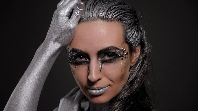 Close-up of a female model with silver skin and hands, with bright makeup with stones posing in the studio on a black background.