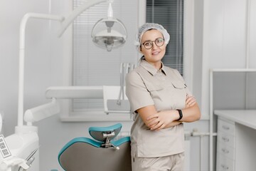Obraz premium Beautiful dentist woman in glasses and a medical cap stands with her arms crossed on her chest in a dental clinic. Concept of painless dental treatment