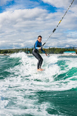 Female in wetsuit riding the waves using of tow rope behind a boat on sunny day. Watersport concept. Young athletic woman learning wakesurfing and perfecting tricks