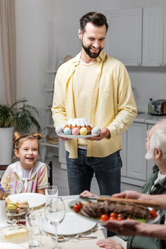 smiling man holding easter cake and painted eggs near family during festive dinner.