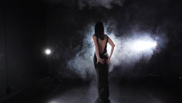 Slender woman in black evening dress with bare back dances on stage in the smoke at night