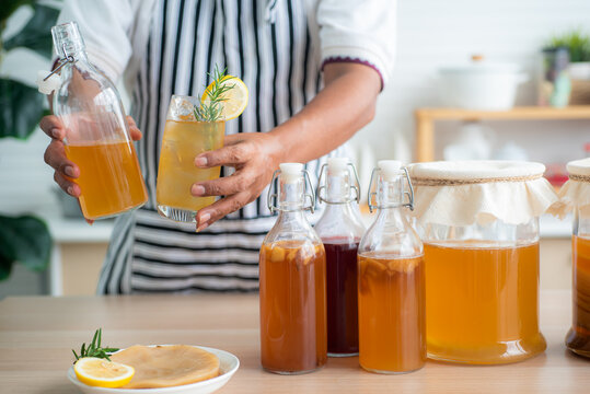 Chef's hand holds a bottle and a glass of Homemade fermented kombucha tea, variety of flavors in bottles and glass jars arranged mix with fruit, scoby on wooden table. Healthy natural probiotic drink.