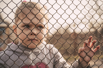 A little refugee girl with a sad look behind a metal fence. The social problem of refugees and...