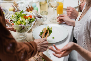 cropped view of woman holding bowl with fresh vegetable salad near blurred family having easter dinner.