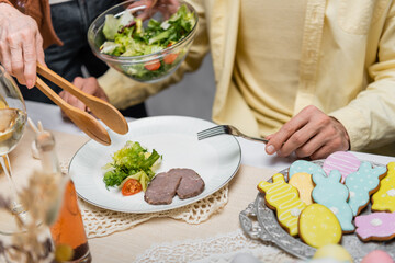 partial view of senior woman holding serving forceps and vegetable salad near adult son.