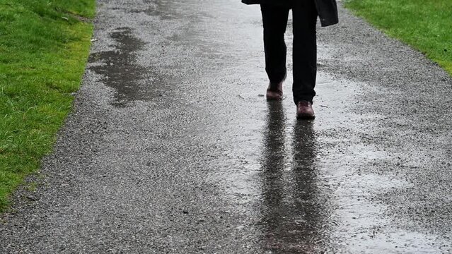 Woman In Black Raincoat And Black Pants Walking On A Gravel Path In A Park On A Wet, Rainy Winter Day, With Standing Rainwater With Grass Lawn 
