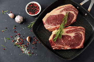 Raw meat, two beef steaks in a grill pan with rosemary and spices on a black background, top view
