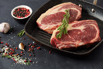 Raw meat, two beef steaks in a grill pan with rosemary and spices on a black background, top view