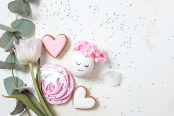 Stylishly decorated egg with pink flowers and sweets on a white background. Creative card for Easter. Romantic egg with a sprig of eucalyptus and desserts view from above