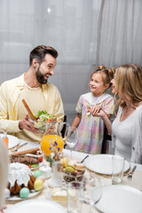 excited man holding bowl with vegetable salad near daughter and wife during easter dinner.