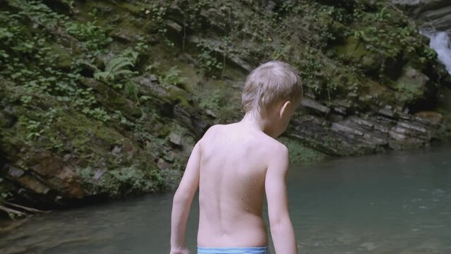 The boy enters the water near the waterfall to swim. CREATIVE. A white child in blue shorts enters the water. In the mountains there is a guy waist-deep in water