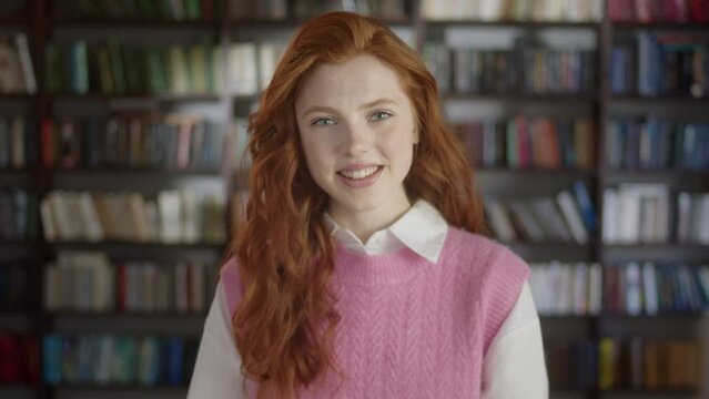 Portrait of Red-haired Young Woman Student Standing in Front of the Camera and Laughing in High School Library. Education, Literature and People Concept.