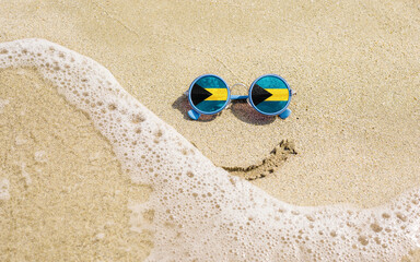 Fototapeta na wymiar Sunglasses with flag of Bahamas on a sandy beach. Nearby is a sea lightning and a painted smile. The concept of a successful vacation in the resorts of Bahamas.