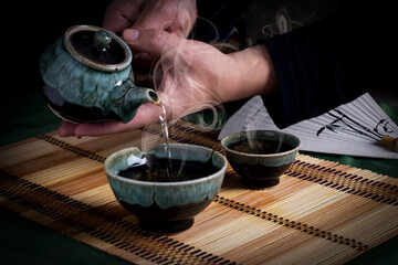 Pouring hot tea from a teapot into a glass on the mat covered table on a dark wooden background.