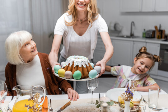 smiling woman holding easter cake with painted eggs near mother and daughter.