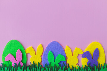 Easter hunt concept. Colorful Easter bunnies and eggs in grass on pink background, top view with space for text. Happy Easter! Pink, yellow, purple artificial bunny and egg decor.
