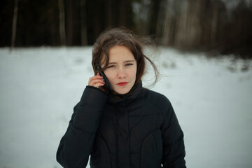 Girl is talking on phone in woods. Schoolgirl is alone in park in winter. Woman holds smartphone near her head. Walk in cold weather.