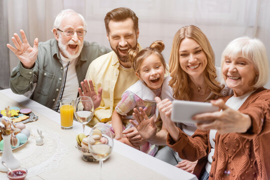 senior woman holding smartphone near excited family waving hands during easter dinner.