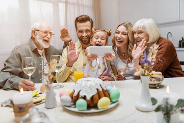 cheerful family waving hands during video call on smartphone near table with easter dinner.