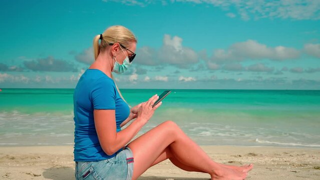 girl in a coronavirus mask sit on a sandy beach and look at the screen of a smartphone on the blue sea.