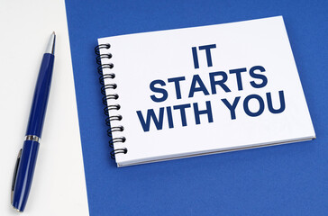 On a blue-white surface lies a pen and a notebook with the inscription - It Starts With You
