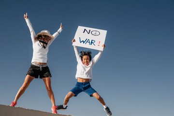 a Caucasian mother in a white hat, jumps next to her Asian son holding a sign that reads "no war". concept war and peace. conflict and protest. diversity.