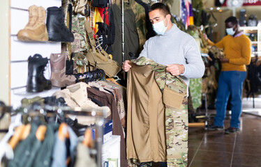 Adult man in protective mask chooses camouflage uniform at army store