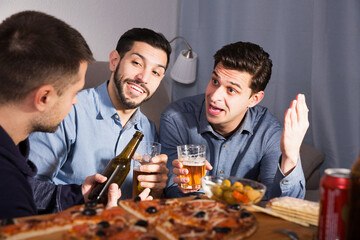 Three adult cheerful men talking and laughing while enjoying beer and pizza at home